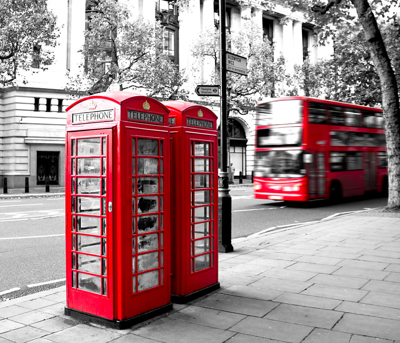 Black and White photo of a London road, with two British red telephone boxes and a red double-decker bus in colour.